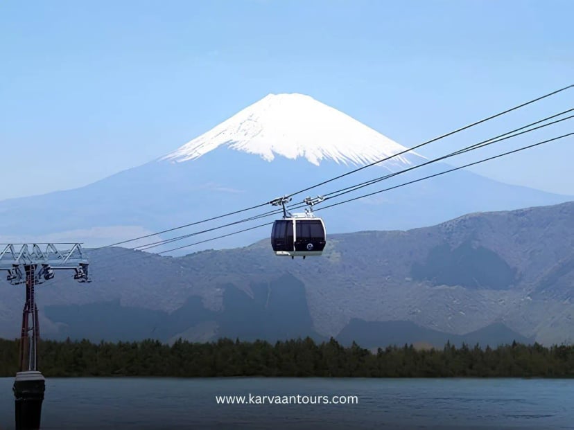 A cable car suspended on wires above the water of Lake Kawaguchiko, with the iconic, snow-covered peak of Mount Fuji dominating the background under a clear, bright sky.