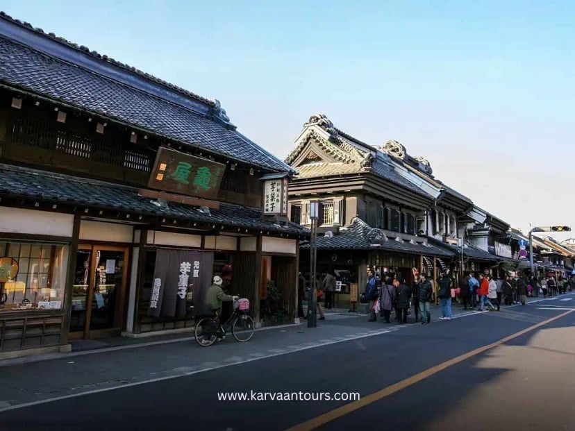 A narrow, cobblestone alleyway in Kawagoe lined with traditional wooden buildings, including a charming cafe with a circular sign, with a person standing by the entrance.