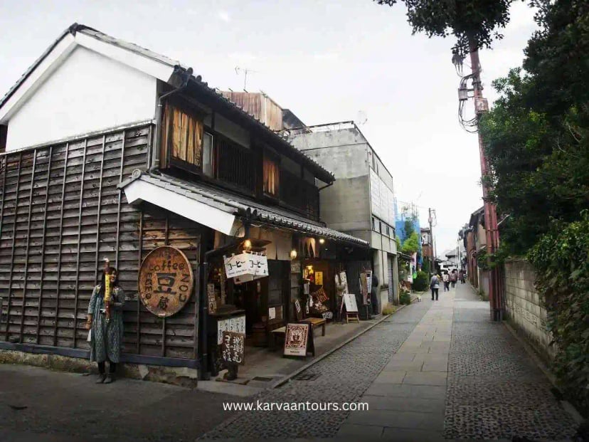 A street scene in Kawagoe’s historic Kurazukuri district, featuring traditional merchant houses with dark tiled roofs and earthen walls, with pedestrians walking along the sidewalk and a person riding a bicycle in the foreground.