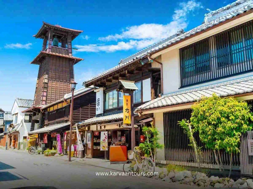 A picturesque view of the iconic Toki no Kane (Bell Tower) in Kawagoe, rising above traditional wooden storefronts and historic architecture under a bright, clear blue sky.