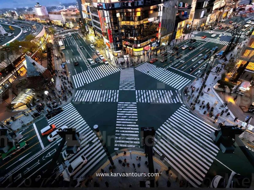 An aerial, high-angle perspective looking directly down at the famous Shibuya Crossing in Tokyo, showing the distinct white pedestrian crosswalks surrounded by city streets and buildings.