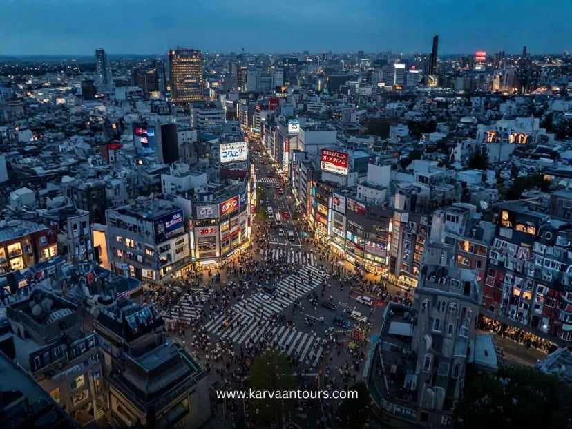 A wide-angle, elevated nighttime aerial shot of the busy Shibuya Crossing, glowing with vibrant neon signs and bustling city lights as crowds move through the iconic intersection.