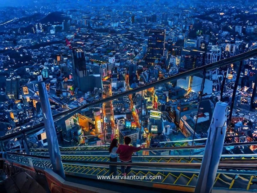 A breathtaking night view from the Shibuya Sky observation deck, where a couple stands on an outdoor escalator looking out over the sprawling, illuminated Tokyo cityscape.
