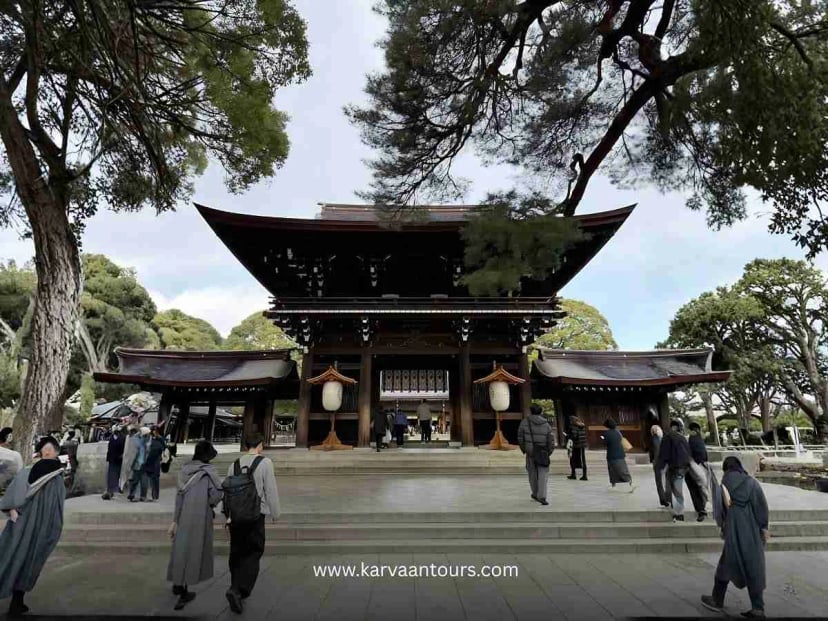 A view of the impressive main hall of Meiji Jingu Shrine in Tokyo, featuring traditional wooden architecture, large white hanging lanterns, and visitors walking through the courtyard under the shade of mature trees.