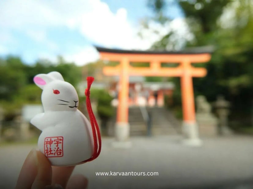 A close-up shot of a small, white ceramic rabbit omamori (amulet) with red accents, held in a hand with a blurred traditional red torii gate of a shrine visible in the background.