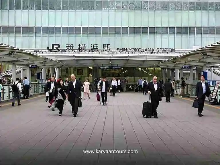A busy daytime scene at the JR entrance of Shin-Yokohama Station, showing commuters and travelers walking through the bustling transit hub.