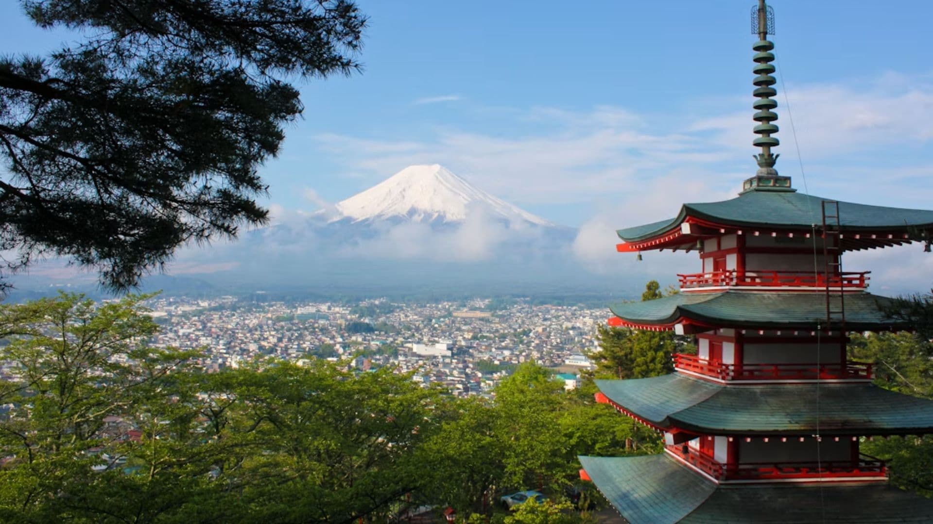 A traditional red and white five-story pagoda framed by trees overlooking a city and Mount Fuji.