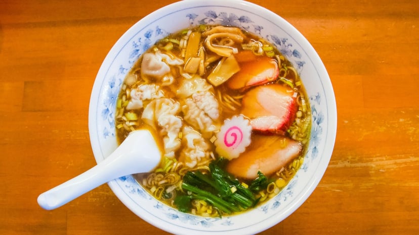 Top view of a fresh bowl of Japanese ramen broth with wontons, sliced chashu pork, narutomaki fish cake, bamboo shoots, and green vegetables.