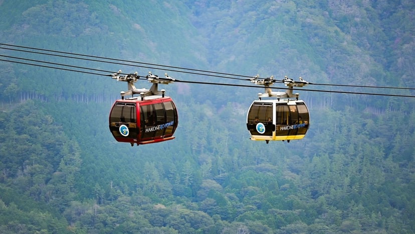 Red and yellow Swiss-style gondola cabins gliding along steel cables high above lush green forested mountains.