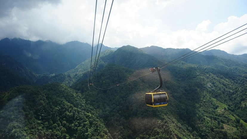 First-person POV looking down from a high-altitude cable car cabin suspended over dense green mountain slopes and valley ridges.