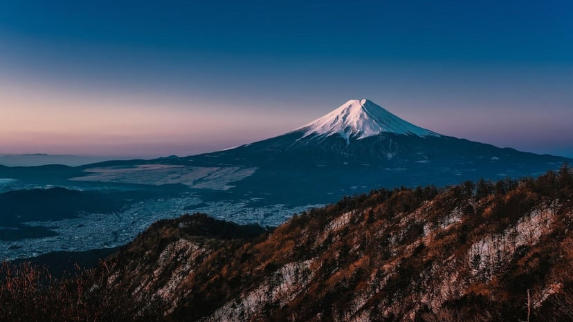 Clear panoramic view of snow-capped Mount Fuji rising above dark mountain ridges and surrounding valley under a twilight sky.