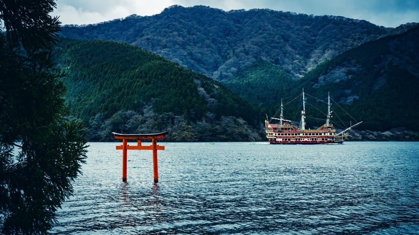 Red pirate sightseeing ship sailing across Lake Ashi near the floating vermilion Torii gate of Hakone Shrine against lush green mountains.